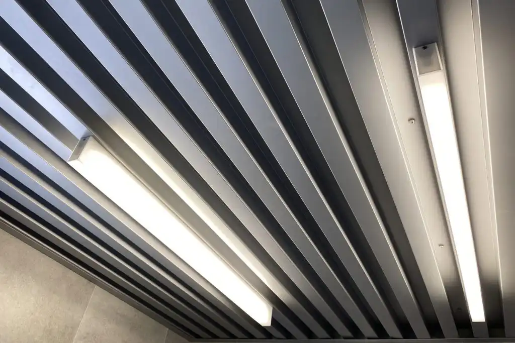 Podhled z plechového ohýbaného profilu DEKPROFILE vyrobeného na míru, doplněný o stropní osvětlení – moderní kovový designový podhled pro architektonické interiéry.